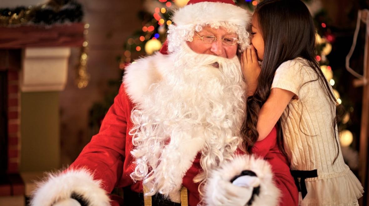 A girl in a white dress with long brown hair whispers in Santa's ear Best Santas for Santa photos around Seattle, Eastside and South Sound
